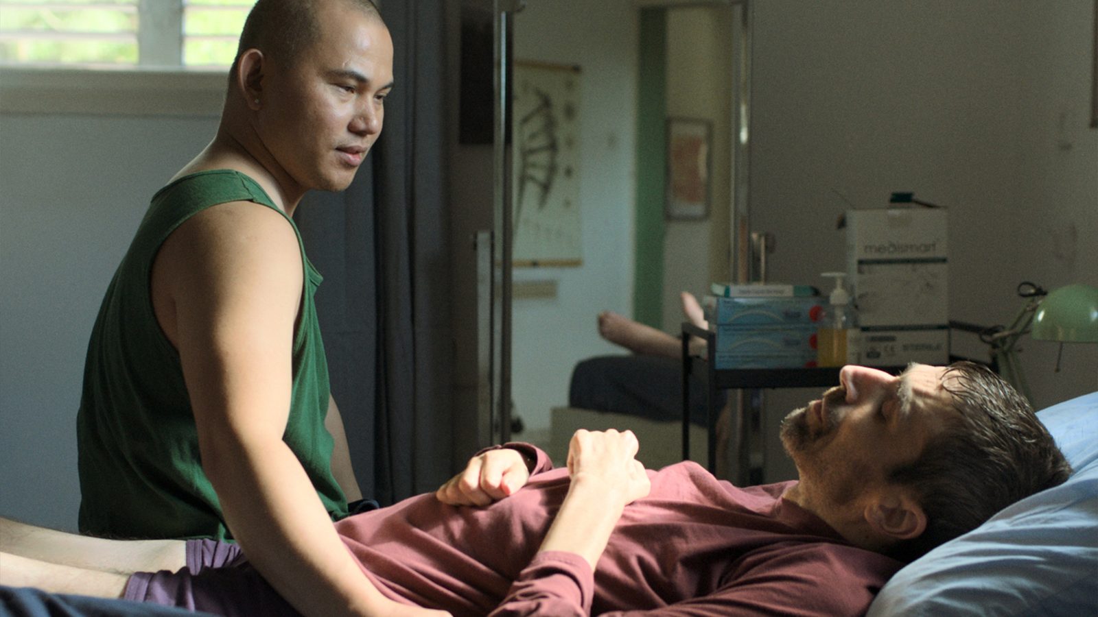 Oliver sits beside Mickey, holding his hand, in a still from Another Body